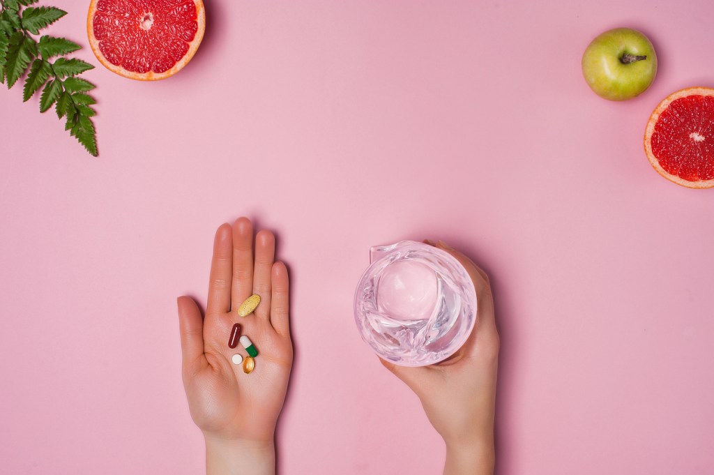 Vitamins and a glass of water in female hands on a pink background. Place for text. Grapefruit, apple and green leaf on the background. Healthy lifestyle concept. Top view